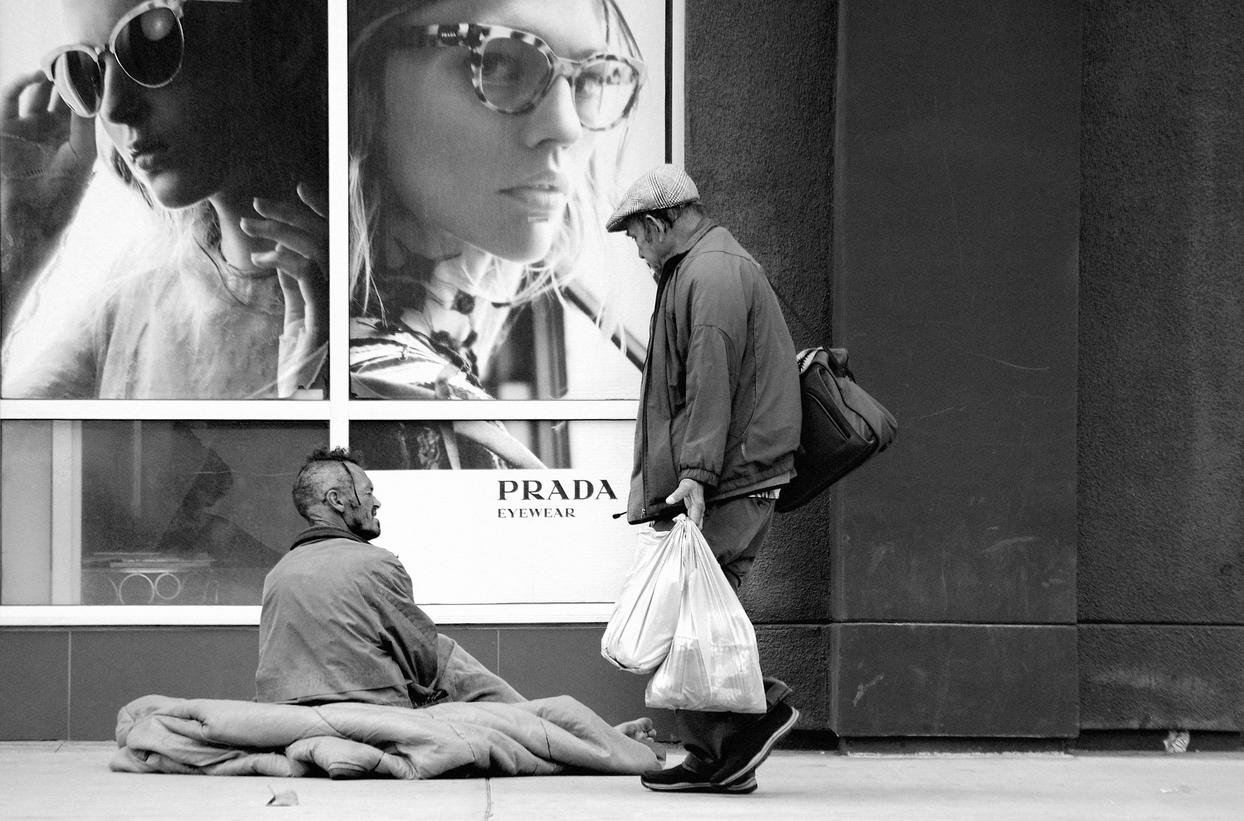Homeless Person and Pedestrian in front of fancy Prada store front.